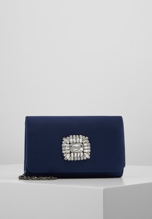 Pochette bleu marine avec une décoration rectangulaire en pierre précieuse argentée centrée sur le devant, posée sur une plateforme blanche avec une chaîne argentée.