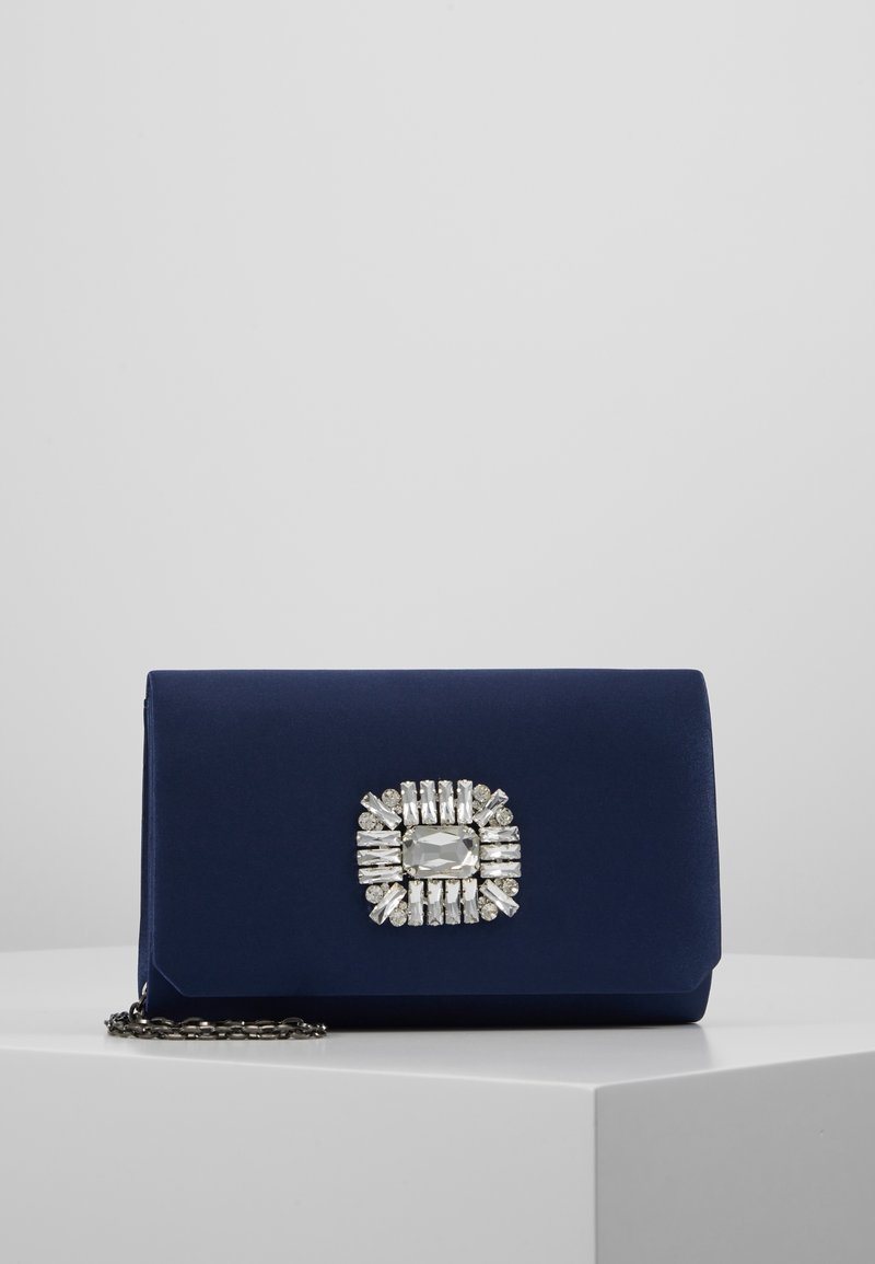 Pochette bleu marine avec une décoration rectangulaire en pierre précieuse argentée centrée sur le devant, posée sur une plateforme blanche avec une chaîne argentée.