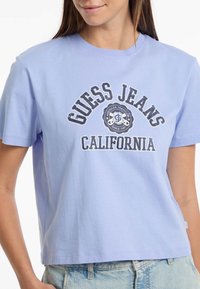 Šviesiai mėlynas medvilninis marškinėlis su ryškiu juodu ir baltu "GUESS JEANS CALIFORNIA" grafiniu raštu bei klasikinio apvalaus kaklo dizainu.