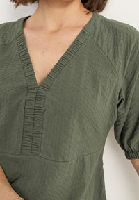 Zielona, teksturowana bluzka z dekoltem w kształcie V i marszczeniami wzdłuż dekoltu i rękawów, z krótkimi bufiastymi rękawami.