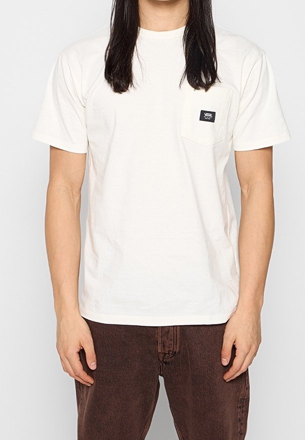 Personne aux longs cheveux foncés portant un t-shirt blanc uni à manches courtes avec un petit logo noir sur la poche poitrine et un pantalon marron foncé.