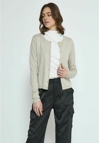 Cardigan beige chiaro con scollo rotondo, maniche lunghe e chiusura con bottoni. Abbinato a pantaloni cargo neri, con tasche e texture liscia.
