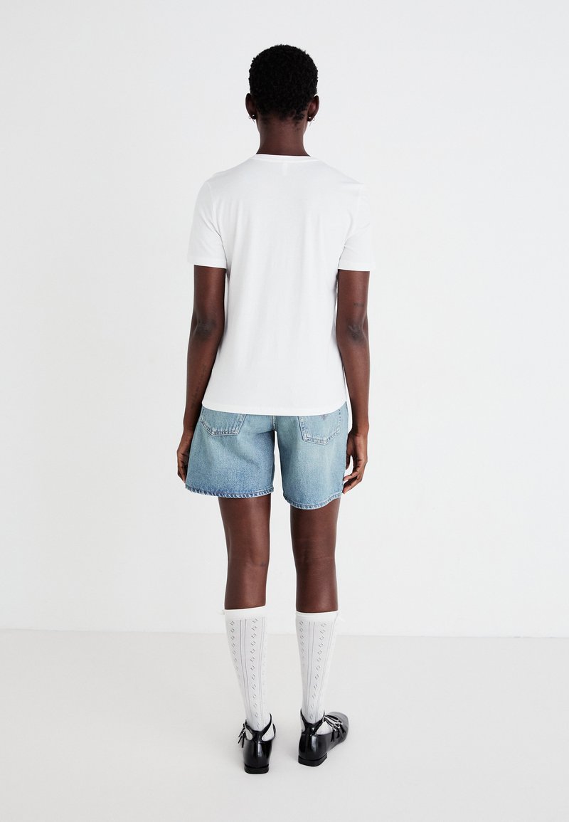 T-shirt blanc à manches courtes, shorts en denim bleu clair, chaussettes blanches montantes à motif, et chaussures noires vues de derrière.