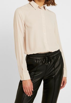 Woman wearing a light beige button-up blouse tucked into high-waisted black faux leather pants with a drawstring waist.