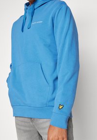 Felpa blu con cappuccio regolabile tramite coulisse, tasca frontale a marsupio e polsini a coste. Presenta un logo ricamato e un accento giallo sulla manica.