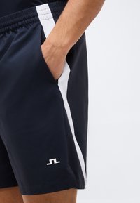 Άνδρας που φορά μπλε σκούρο σορτς με λευκές ρίγες στο πλάι και μικρό λευκό λογότυπο, το χέρι του ακουμπάει στην δεξιά τσέπη.