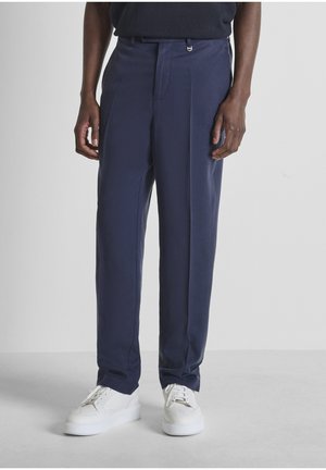 Uomo con pantaloni sartoriali blu navy e sneakers bianche, in piedi contro uno sfondo grigio chiaro.