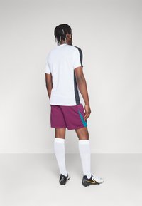 Abbigliamento sportivo composto da una maglietta bianca a maniche corte con pannelli laterali neri e pantaloncini bordeaux con dettagli turchesi, calzini bianchi e scarpini neri.