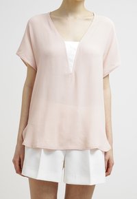 Blouse rose clair à coupe ample, manches courtes et décolleté en V profond. Tissu doux et fluide avec une texture lisse, porté sur un top blanc et un short blanc.