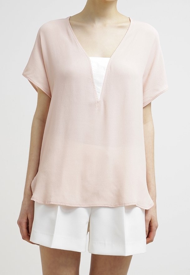 Blouse rose clair à coupe ample, manches courtes et décolleté en V profond. Tissu doux et fluide avec une texture lisse, porté sur un top blanc et un short blanc.