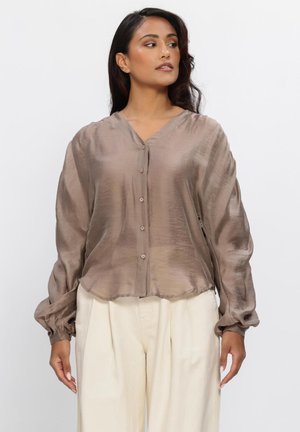 Vrouw draagt een losse, glanzende taupekleurige blouse met knopen, met geplooide mouwen en een broek met hoge taille en wijde pijpen in crèmekleur, afgebeeld tegen een effen achtergrond.