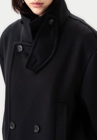 Svart dubbelknäppt rock i mjukt tyg med stora knappar, trattkrage och sidoficka, i en ren, skräddad design.