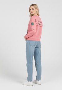 Felpa rosa con stampe grafiche sul fronte e sul retro, abbinata a jeans azzurri con trama leggera e sneakers bianche su uno sfondo neutro.