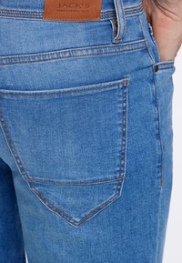 Vaaleansiniset farkut, joissa on ruskea nahkavanne ja erottuva ompelu. Mukana on kaareva takataskumuotoilu ja slim fit -silhuetti.