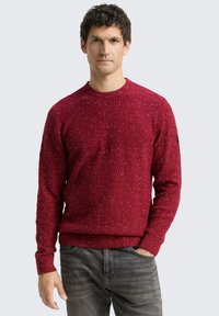 Pull rouge avec un motif tricoté texturé, encolure ronde et poignets et ourlet côtelés. Associé à un jean gris.