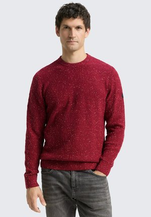 Roter Pullover mit strukturiertem Strickmuster, runder Ausschnitt und gerippten Bündchen und Saum. Kombiniert mit grauen Jeans.