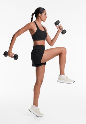 Černá sportovní podprsenka, černé šortky a světlé tenisky. Modelka drží činky a předvádí dynamickou cvičební pozici na bílém pozadí.