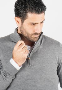 Pull en tricot gris avec un design à fermeture éclair quart de longueur. Poignets et ourlet côtelés. Col de chemise clair visible en dessous. Texture lisse et coupe ajustée.