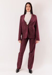 Completo in velluto a coste bordeaux con blazer a un solo bottone e pantaloni su misura. Presenta due tasche frontali e tessuto strutturato. Scarpe nere con punta appuntita.