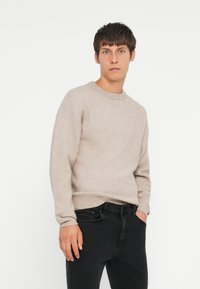 Suéter de punto beige claro con cuello redondo, puños y dobladillo acanalados. Llevado con jeans negros. Textura suave, diseño casual.