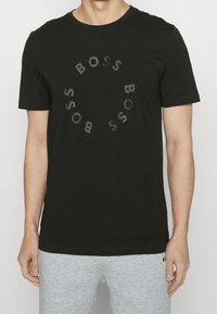 Man i svart kortärmad T-shirt med cirkulärt "BOSS" textmönster på framsidan, kombinerad med ljusgrå avslappnade byxor.