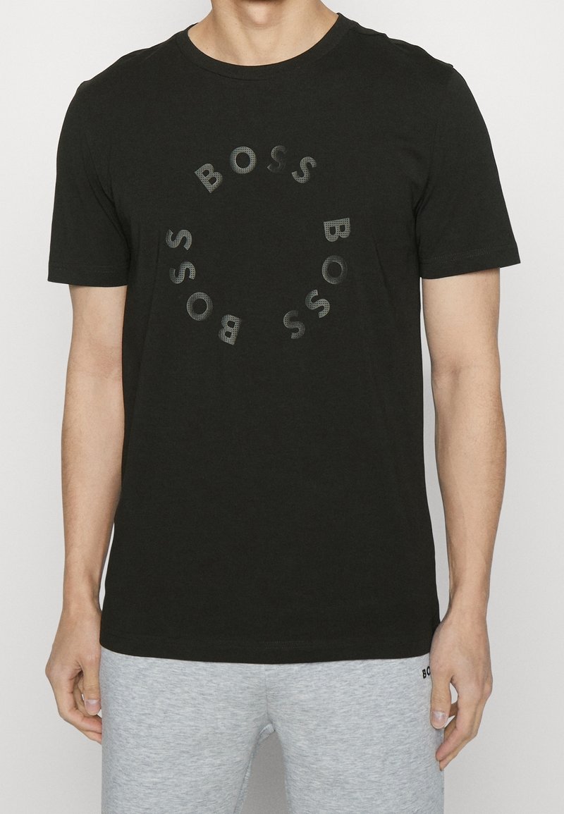 Man i svart kortärmad T-shirt med cirkulärt "BOSS" textmönster på framsidan, kombinerad med ljusgrå avslappnade byxor.