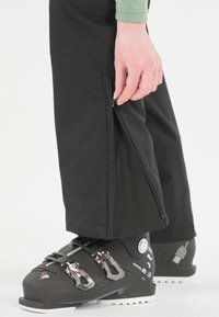 Schwarze Skihosen mit Reißverschlüssen an den Knöcheln, die eine glatte Textur aufweisen. Getragen über schwarzen Skischuhen mit metallischen Schnallen und einem eleganten Design.