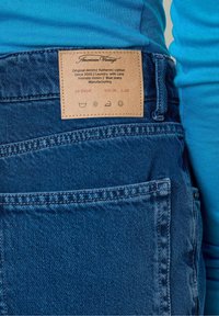 Blauwe denim jeans met een leren label met merknaam, onderhoudsinstructies en maat. Stiksels zijn zichtbaar op de zakken en de tailleband.