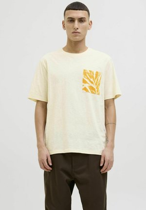T-shirt en coton jaune clair avec une poche patch orange à motifs représentant des feuilles. Manches courtes et encolure ras du cou.