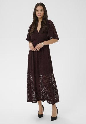 Kobieta o długich, ciemnych włosach, ubrana w fioletową sukienkę midi z koronką oraz czarne szpilki, stojąca z delikatnie złączonymi rękami.