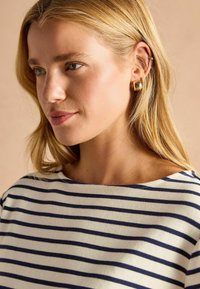 Röstripeeritud puuvillane topp, millel on lai kaelus ja tumesinised triibud kreemival taustal, kantud kuldsete rõngaste kõrvarõngastega. Sile tekstuur on nähtav.