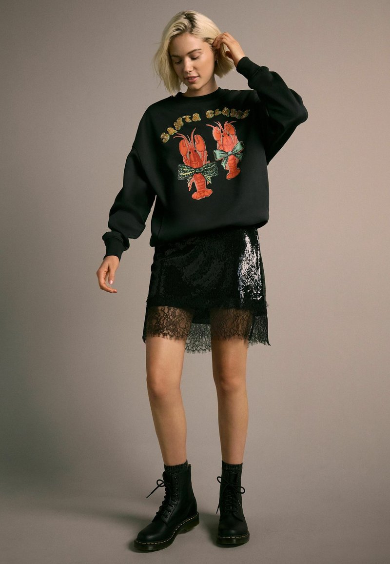 Czarny sweatshirt z grafiką homara, w połączeniu z czarną spódnicą z cekinów z koronkowym wykończeniem. Modelka nosi czarne botki na sznurówki.