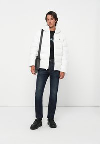 Jonge man in een witte pufferjack, zwart graphic tee, donkere jeans, zwarte schoenen en met een zwarte crossbody tas, staande tegen een witte achtergrond.