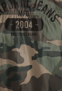 Camo-patterned fabric in shades of green and brown, featuring bold black text "APORAL JEANS MFG 2004 REGISTERED BRAND" printed on it.