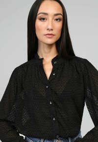 Chemisier noir à boutons avec un tissu texturé, un col plissé et des boutons transparents. Manches longues avec des poignets froncés, apparence semi-transparente.