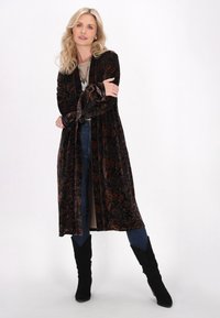 Lunga veste di velluto marrone scuro con un ornato motivo paisley. Presenta maniche ampie e dettagli con lacci. Abbinata a jeans blu e stivali neri.