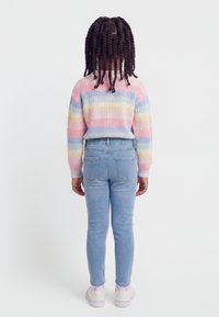 Παιδί με πλεγμένα μαλλιά που φοράει ένα πουλόβερ με παστέλ ρίγες, ανοιχτό μπλε τζιν και λευκά αθλητικά παπούτσια, στέκεται πλάτη σε λευκό φόντο.