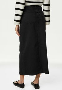 Jupe maxi en denim noir avec une coupe droite, proposant deux poches arrière et une fente latérale discrète. Portée avec des chaussures plates noires.