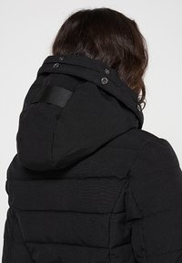 Doudoune noire avec capuche, présentant une texture matelassée, des boutons-pression et un détail de sangle latérale pour ajustement.