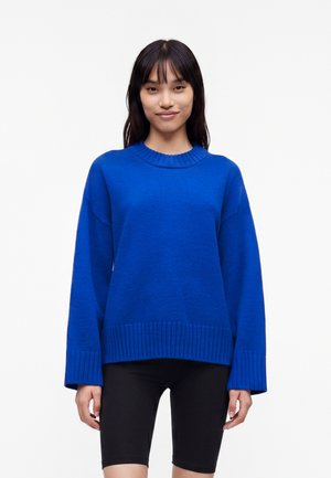 Femme aux cheveux raides et foncés portant un pull oversize bleu vif et un short noir jusqu'aux genoux, debout devant un fond blanc.