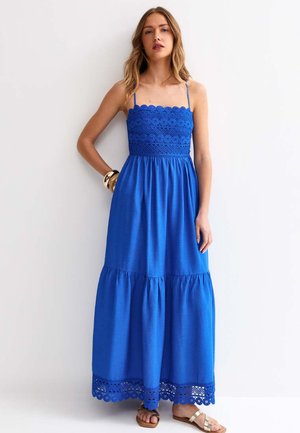 Kobieta ubrana w bezrękawową, królewsko-niebieską sukienkę maxi z szydełkowaną górą i dołem, stoi z jedną ręką na biodrze i sandałami na stopach.