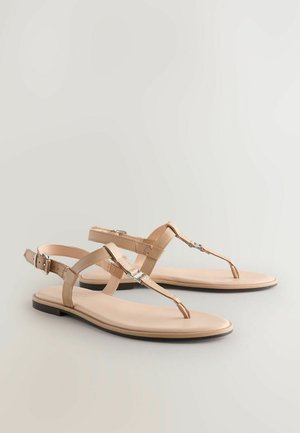 Sandales beiges avec un design à lanière, incluant une lanière en T et une boucle. Matériau lisse semblable au cuir avec une semelle plate et des accents minimalistes.