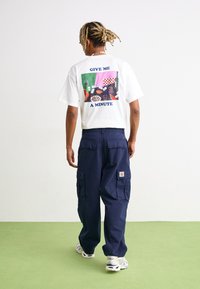 Tricou alb cu mâneci scurte, având un grafism colorat pe spate cu textul "DĂ-MI O MINUTĂ." Purtați cu pantaloni cargo bleu marin.