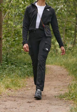 Osoba idąca leśną ścieżką, ubrana w czarną kurtkę, spodnie bojówki, czarne buty trekkingowe, niosąca małą torbę na pasku przewieszoną przez ramię.