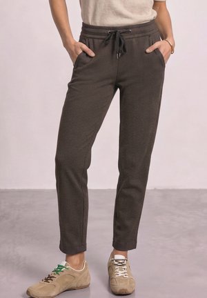 Femme portant un pantalon slim marron foncé avec taille à cordon, mains dans les poches, baskets beige avec lacets dépareillés et un haut beige clair.