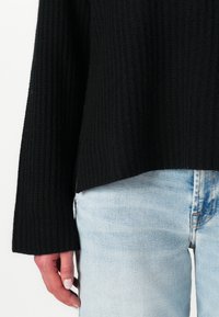 Pull à côtes noir avec manches longues, présentant une coupe légèrement décontractée et un ourlet plus long à l'arrière. Un jeans en denim bleu est porté.