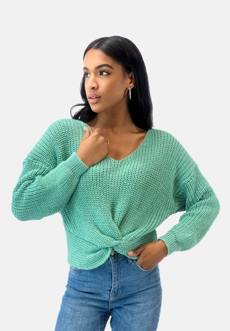 Mintgrüner Strickpullover mit V-Ausschnitt und verdrehtem Frontdetail, kombiniert mit hellblauen Jeans; strukturiertes Gewebe und lockere Passform.