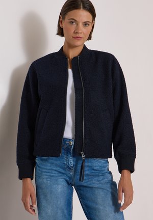 Femme portant une veste zippée texturée bleu marine, chemise blanche et jean bleu, debout sur un fond clair uni.