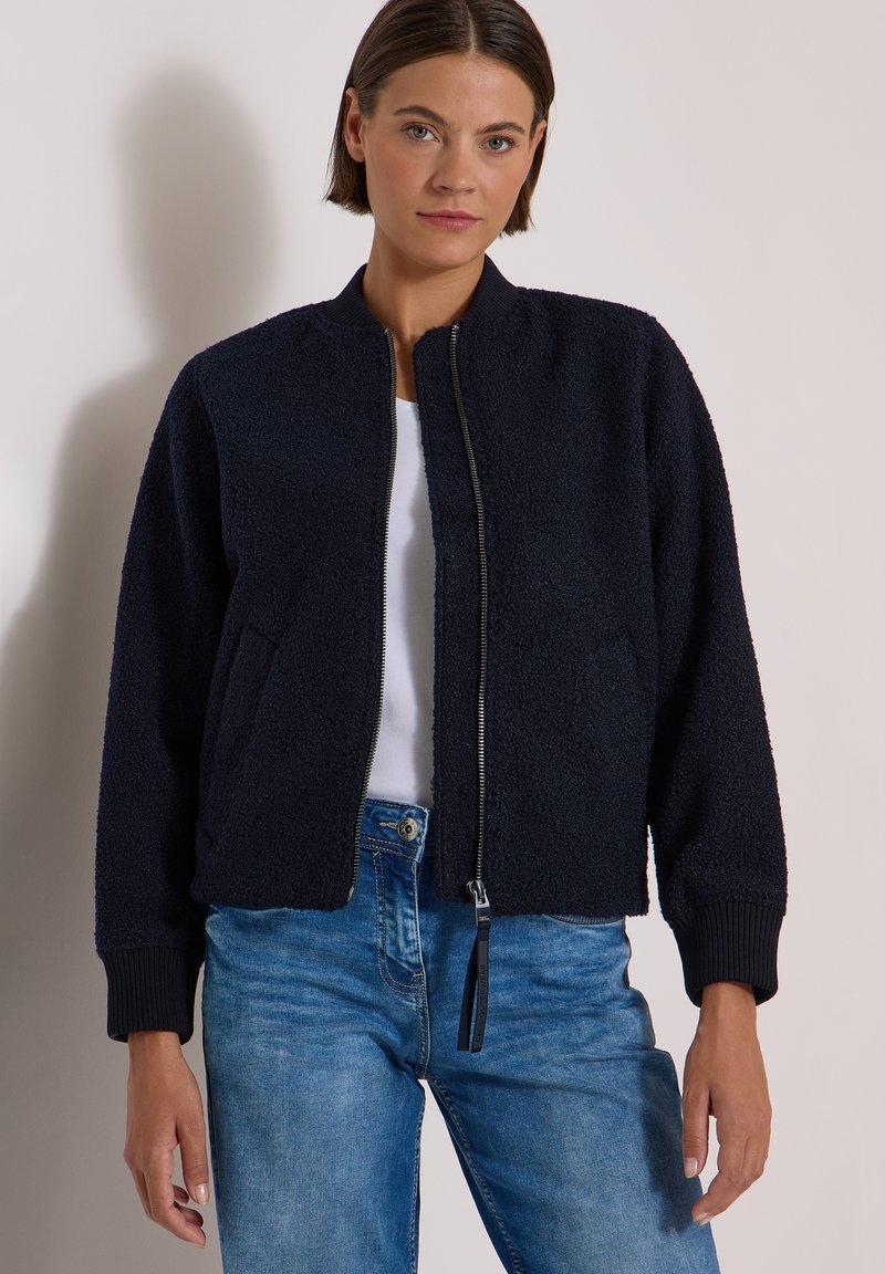 Femme portant une veste zippée texturée bleu marine, chemise blanche et jean bleu, debout sur un fond clair uni.