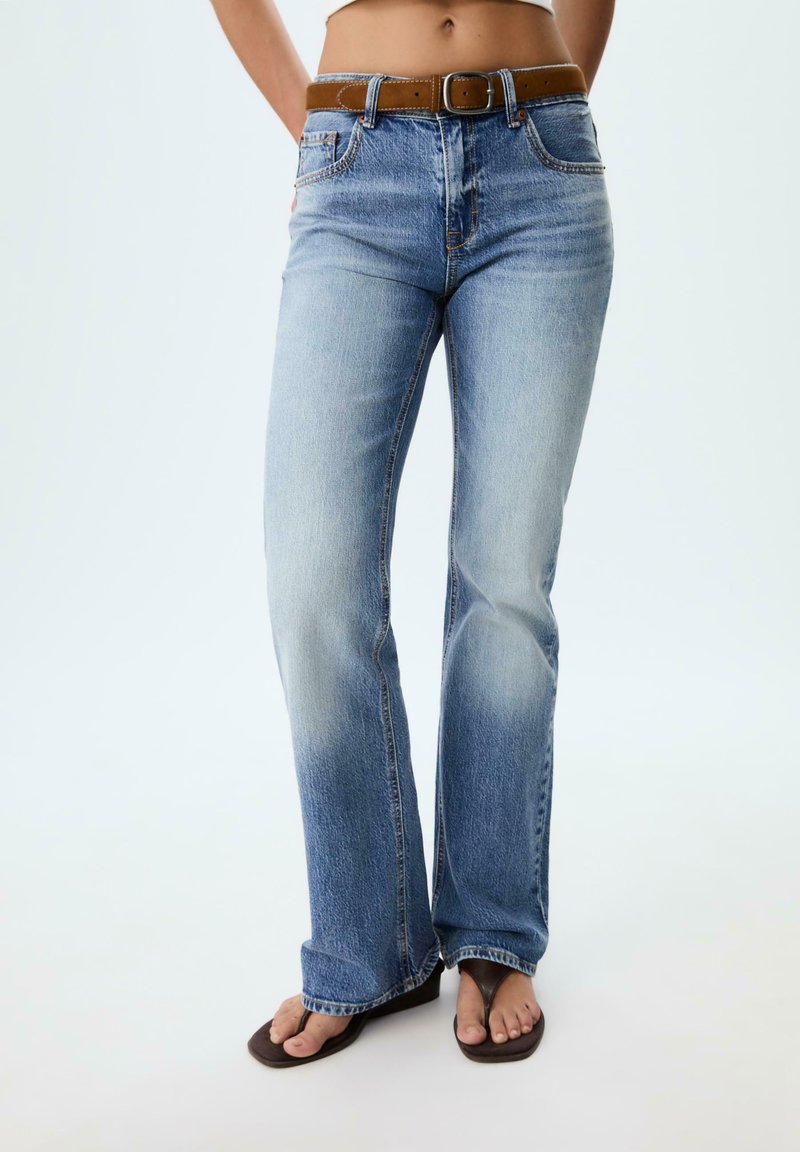 Jean flare bleu délavé avec une teinte claire, design à cinq poches, ceinture en cuir brun, et tongs brunes décontractées.